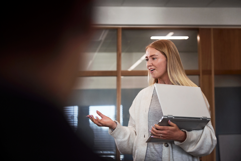 Kvinna med dator som står upp och berättar något för andra i ett konferensrum. 