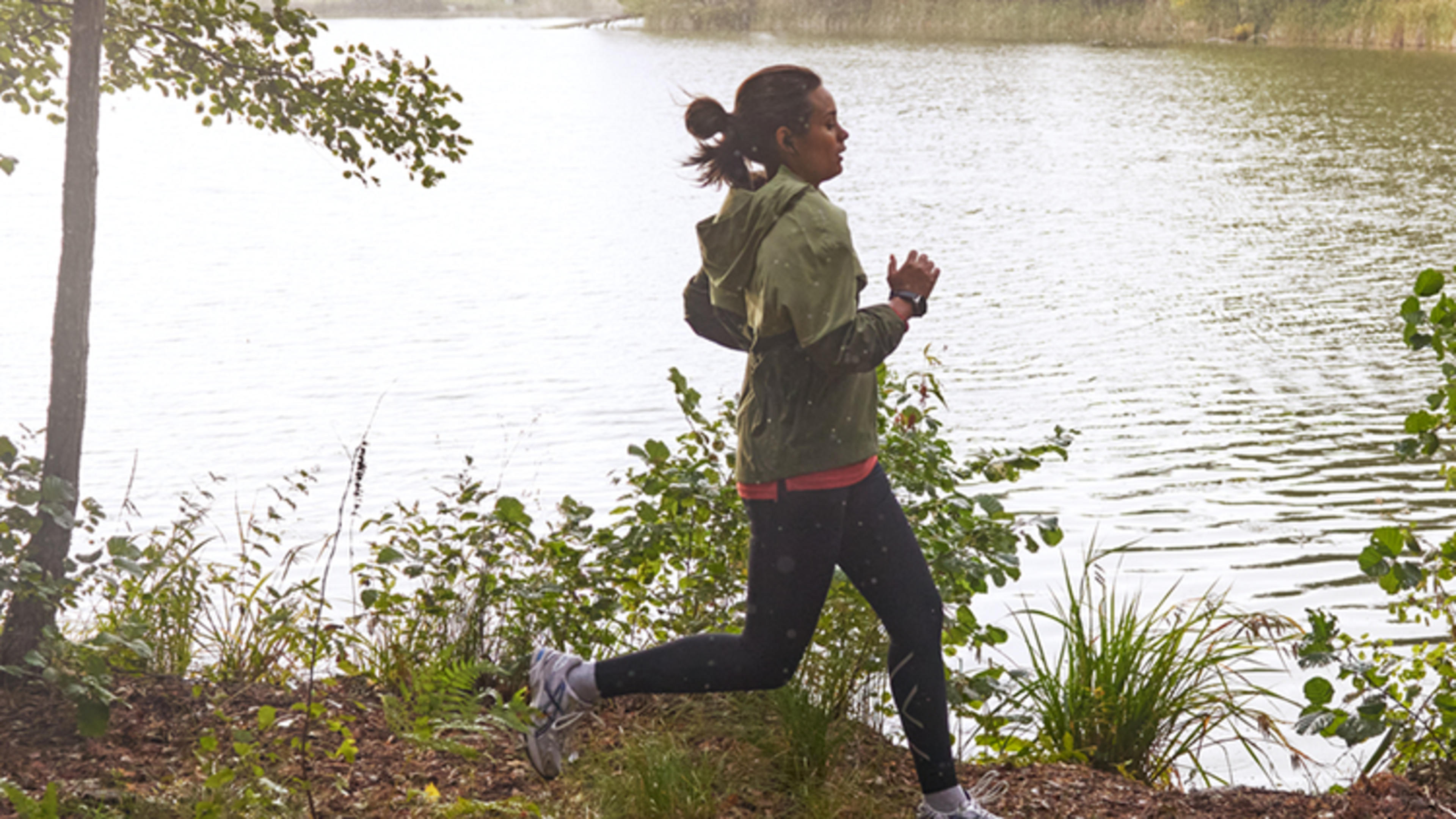 Kvinna joggar i skogen framför sjö.
