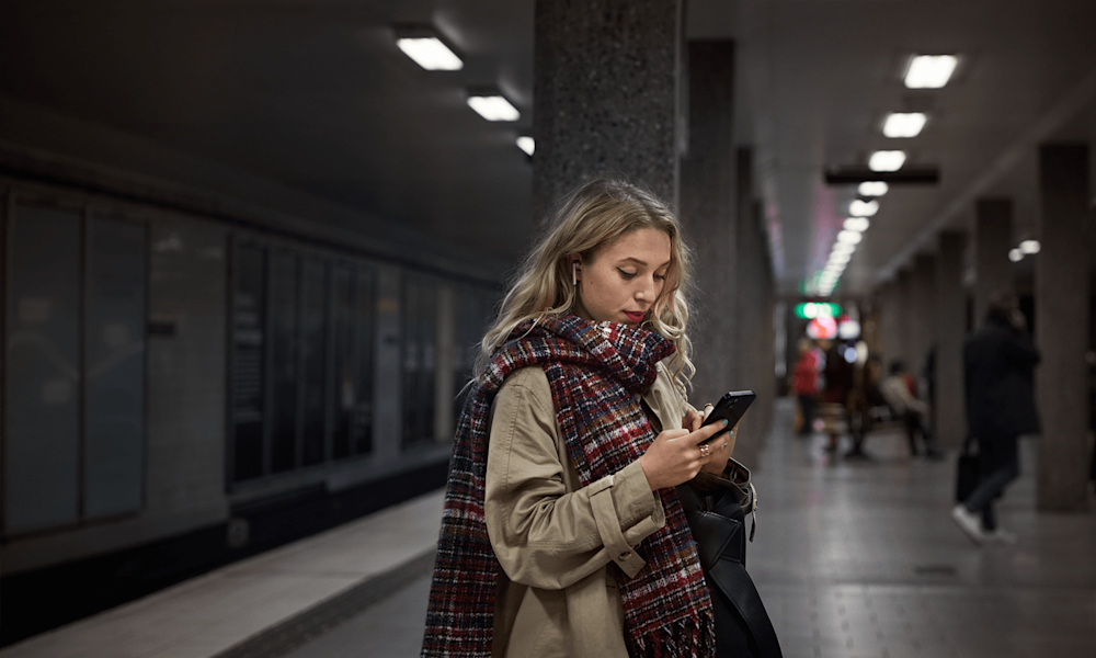 Tjej som står och surfar på sin mobil på en tunnelbaneperrong.