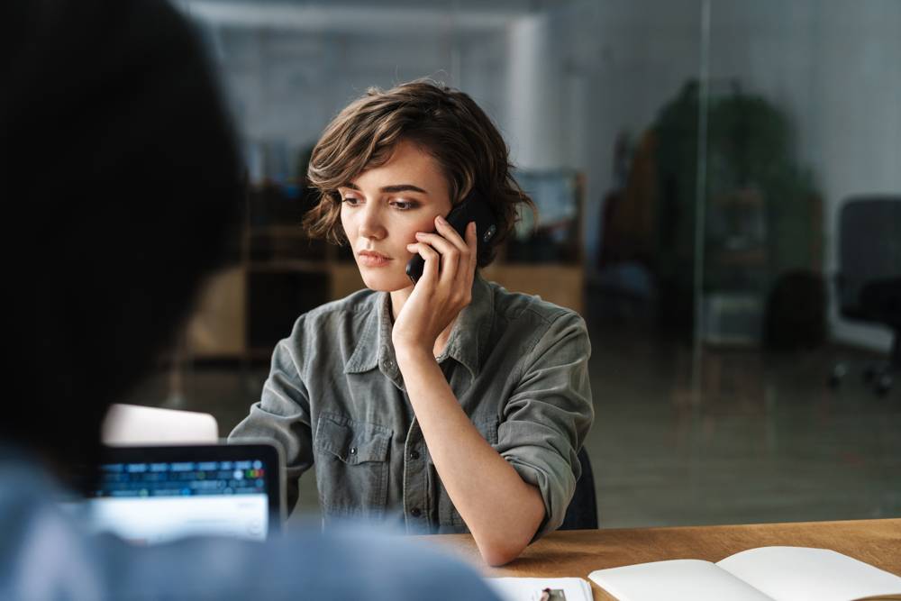 Kvinna pratar i telefon på kontor, orolig, kollega med laptop i förgrunden.