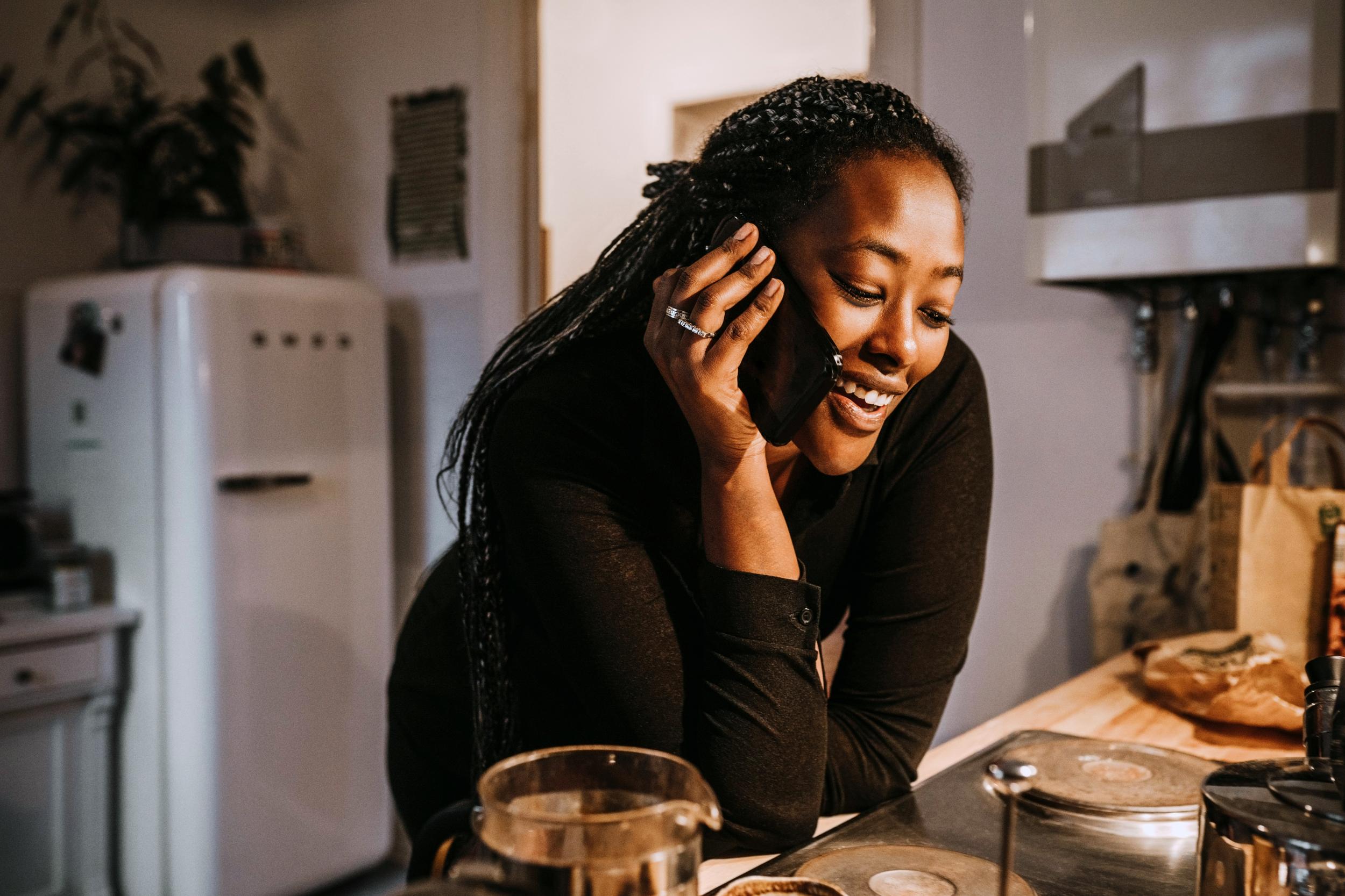 Kvinna som står vid spisen i köket och pratar i sin telefon och ler.