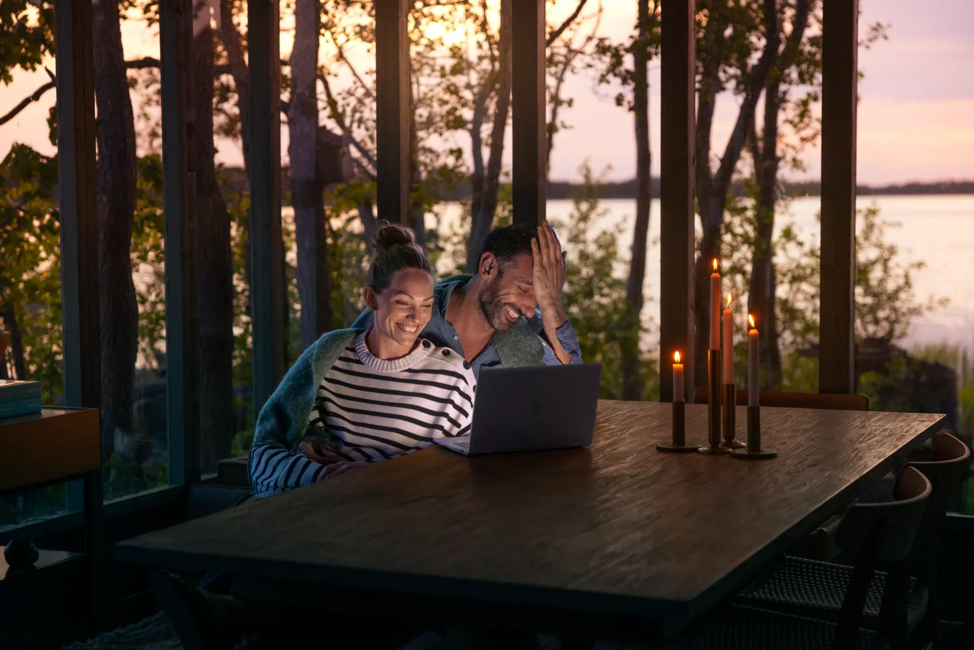 En man och en kvinna sitter vid bordet med levande ljus och tittar på en laptop tillsammans och skrattar med en idyllisk skärgårdsmiljö bakom sig.