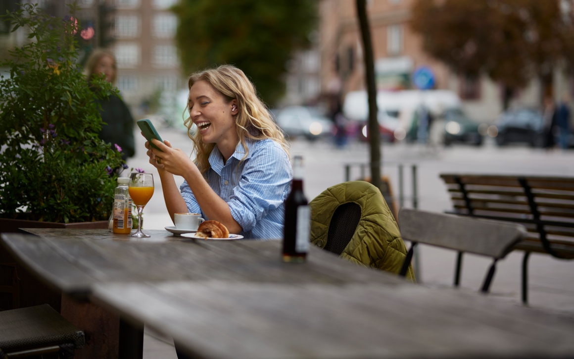 Kvinna på uteservering skrattar med mobil i handen.