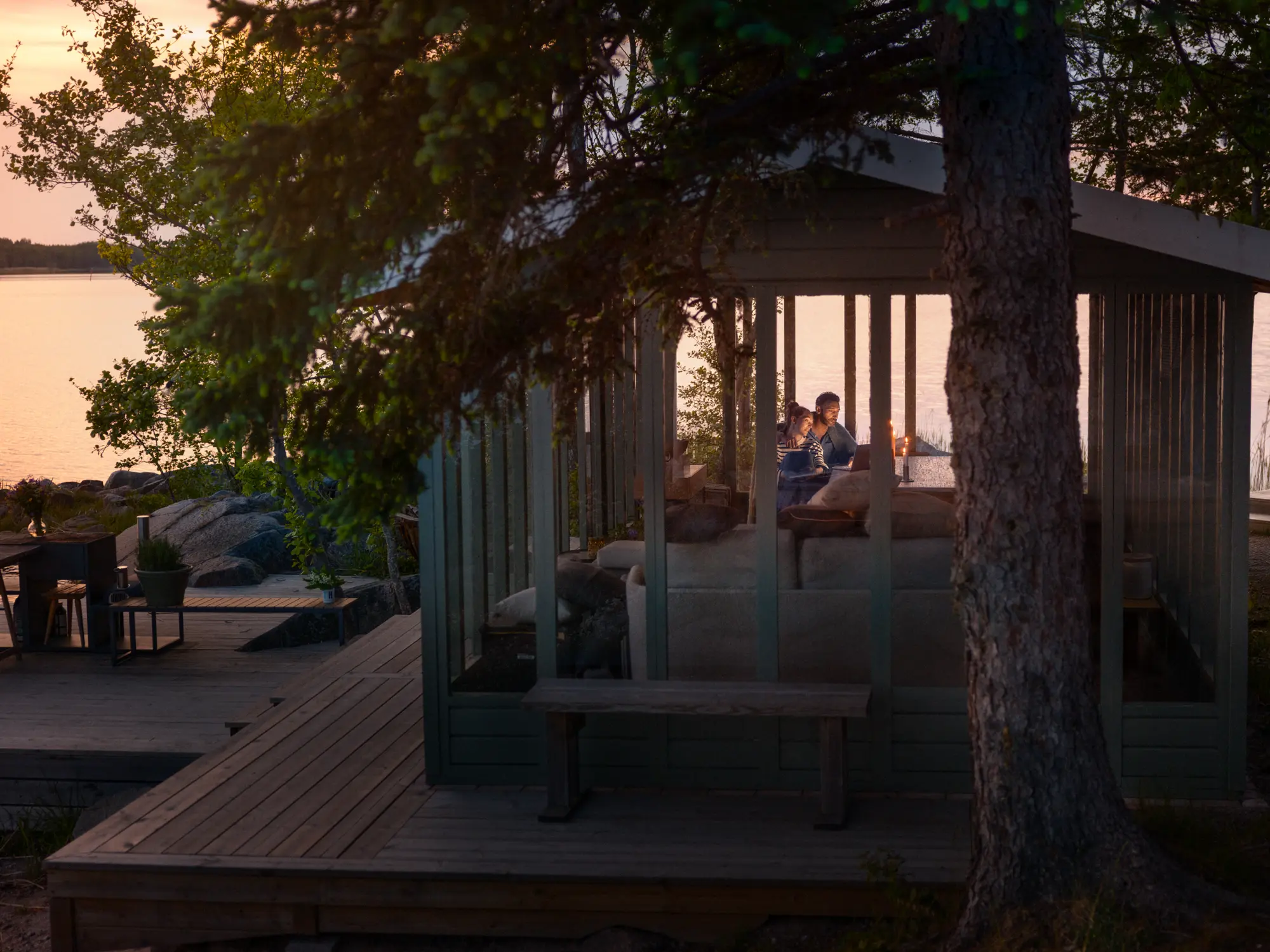 En man och en kvinna som sitter i en mysig stuga precis vid vattnet, som symboliserar Telia Play och Telia tv-paket ger underhållning och bekvämlighet även på avlägsna platser.