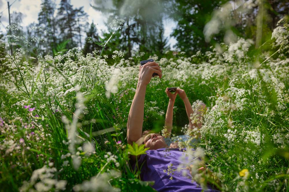 Två ungdomar ligger på rygg i högt gräs och håller upp sina telefoner mot himlen