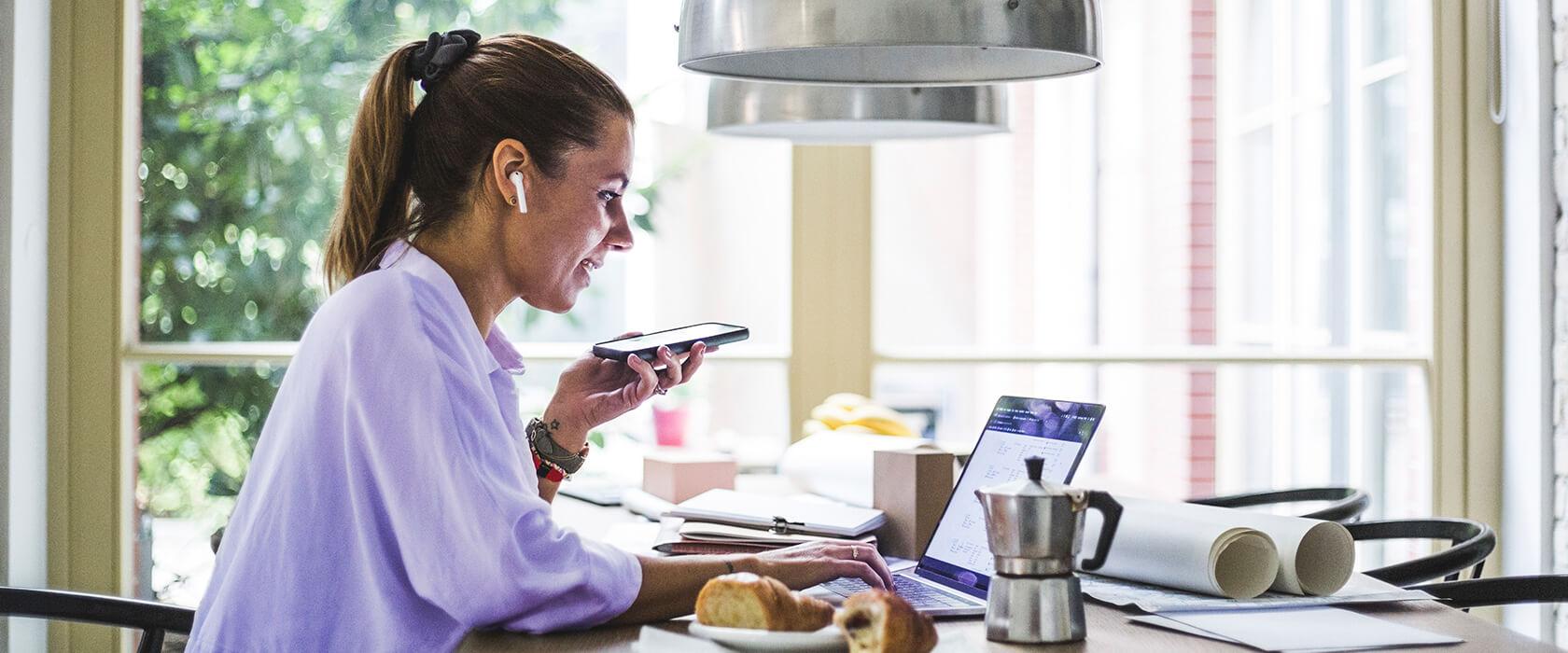 Kvinna med hörlurar som pratar i högtalartelefon och tittar på sin laptop.