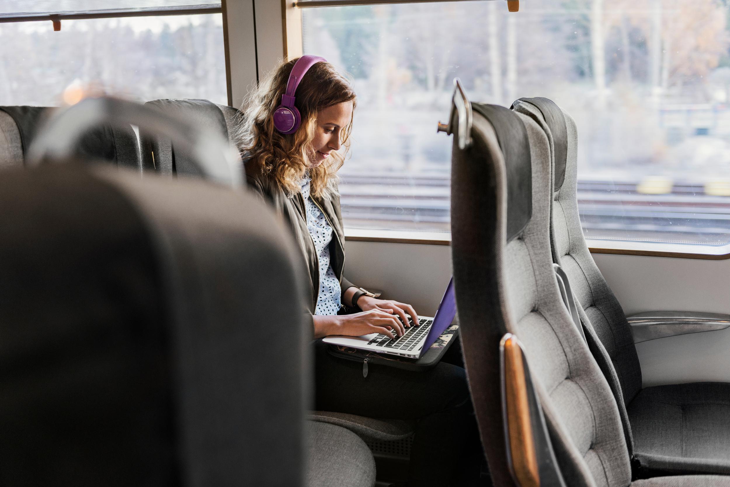En kvinna med hörlurar sitter på ett tåg och jobbar på en laptop.