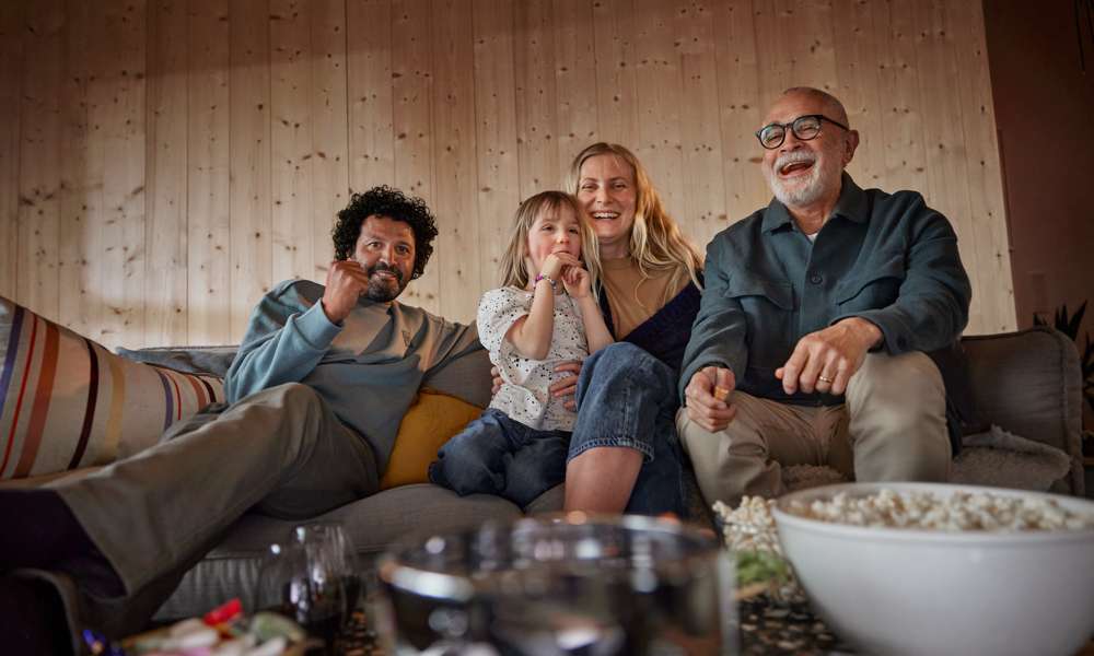 En familj med tre generationer sitter tillsammans i soffan och tittar på tv och skrattar med en stor popcornskål på tv-bordet framför sig.