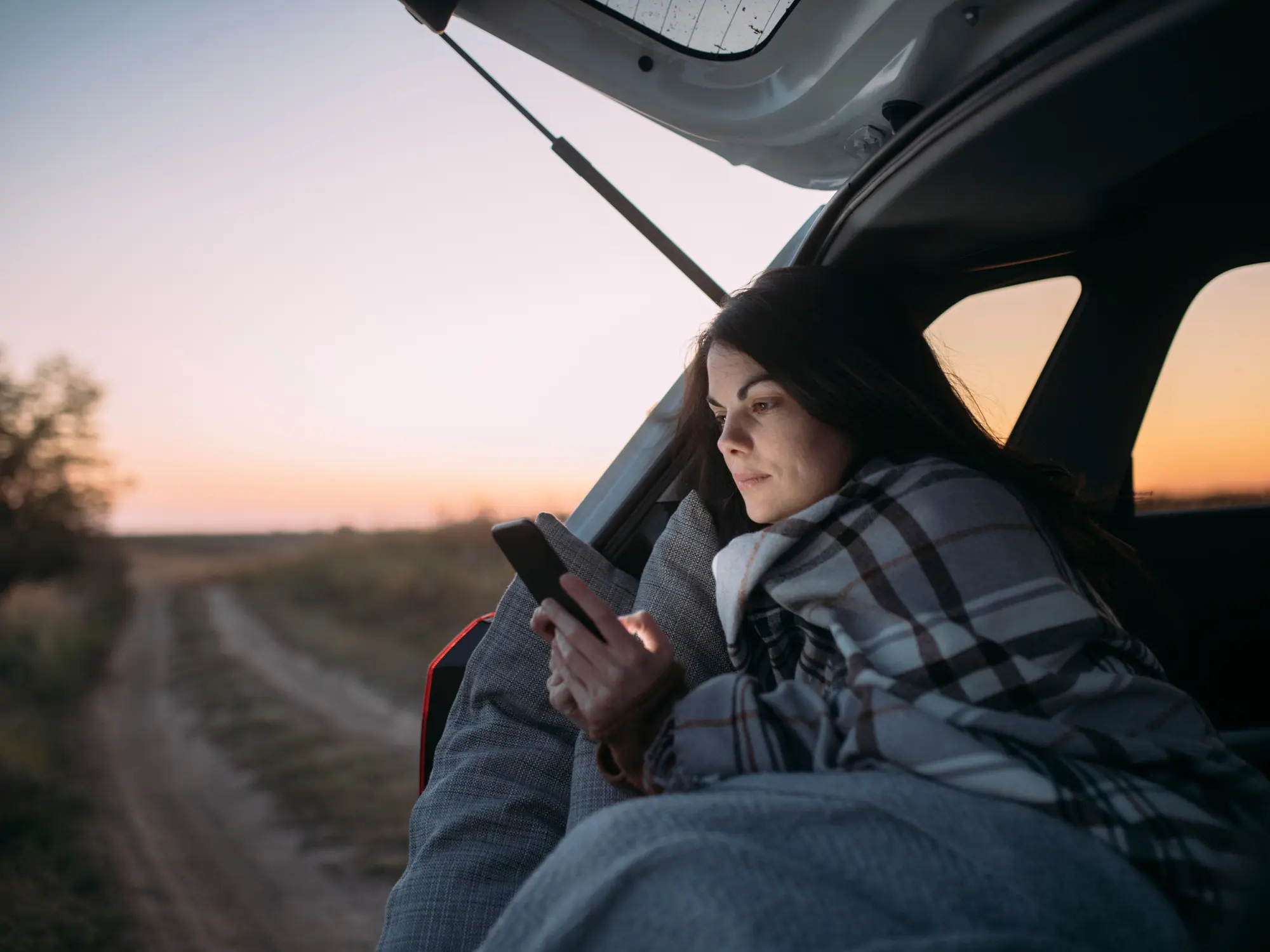 En tjej sitter i bilen en sensommarkväll, omvirad i en filt och tittar på sin mobil med solnedgången bakom sig. 